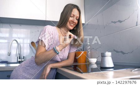 Happy woman is sitting at the kitchen table by the induction hob and brewing coffee in a turk. She admires her jewelry, a pink stone 142297676