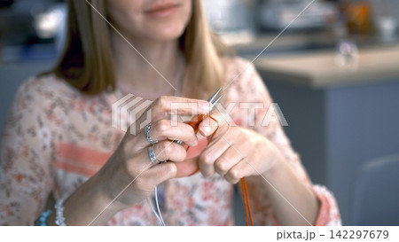 Close-up of a woman knitting a scarf made of orange yarn. Hobbies and creativity. The pleasant pastime of a housewife 142297679