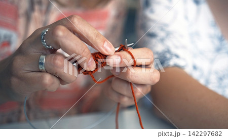 A young specialist knits a scarf made of red and orange yarn, close-up. Art and creativity. Knitting with knitting needles A young specialist knits a scarf made of red and orange yarn, close-up. Art and creativity. Knitting with knitting needles 142297682