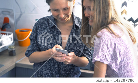 Two girlfriends are standing in the kitchen and watching funny videos on their mobile phones. Lesbians are having fun. They smile and laugh Two girlfriends are standing in the kitchen and watching funny videos on their mobile phones. Lesbians are having fun. They smile and laugh 142297790