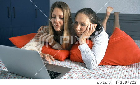 Two lesbian lovers hug each other lying on the bed in front of a laptop. Female students watch movies on their personal computer 142297822