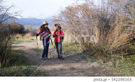 Two tourists are standing on a dirt road and chatting with each other. Two women with backpacks and trekking poles are relaxing after a long hike Two tourists are standing on a dirt road and chatting with each other. Two women with backpacks and trekking poles are relaxing after a long hike 142298103