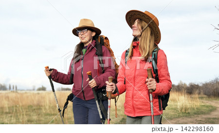 Two happy women in hats and backpacks are walking across the field. Travelers travel together. Outdoor fitness. Unity with nature Two happy women in hats and backpacks are walking across the field. Travelers travel together. Outdoor fitness. Unity with nature 142298160