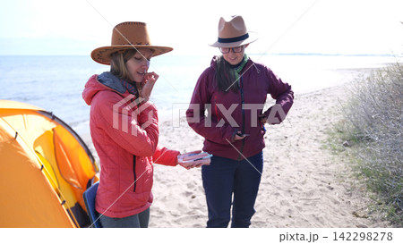 Two funny lesbians are going to roast sausages on a campfire. Two young women are standing on the beach next to a tent. Weekend picnic by the sea Two funny lesbians are going to roast sausages on a campfire. Two young women are standing on the beach next to a tent. Weekend picnic by the sea 142298278