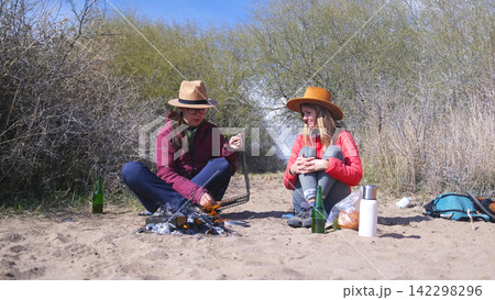 Two beauties are sitting on the beach by the campfire, one of them is frying sausages on a grill. Best friends are relaxing by the sea 142298296
