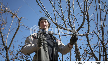 A happy young amateur gardener is working in the garden. A man in a hat and gloves holds a pruner in his hands and prepares to cut the dry branches of a tree A happy young amateur gardener is working in the garden. A man in a hat and gloves holds a pruner in his hands and prepares to cut the dry branches of a tree 142298463