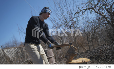 A young student is learning how to chop firewood. Harvesting firewood for the winter. A man is chopping firewood with an axe A young student is learning how to chop firewood. Harvesting firewood for the winter. A man is chopping firewood with an axe 142298476