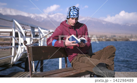 A lonely, handsome student sits on a bench by the water and reads a book against the backdrop of the Kyrgyz mountains. Lake Issyk Kul is the pearl of Central Asia 142298489