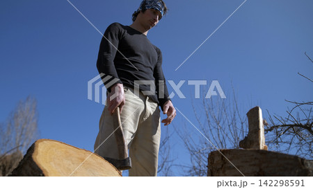 A young man looks at the firewood in dismay, not knowing how to chop it. He holds an axe in his hand in the garden. The sky is clear and blue 142298591
