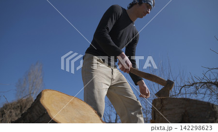 A man is chopping firewood with an axe. The sky is blue and the trees are bare. The man is focused on his task and seems to be enjoying a walk A man is chopping firewood with an axe. The sky is blue and the trees are bare. The man is focused on his task and seems to be enjoying a walk 142298623