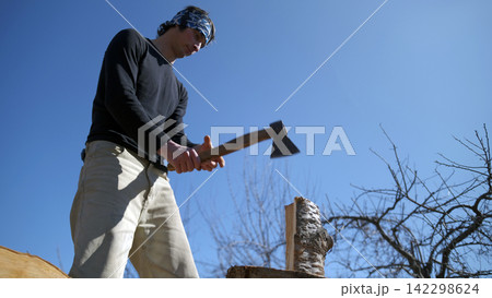 The process of harvesting firewood for the winter outdoors. A man is chopping firewood with an axe. The sky is blue, cloudless and clear The process of harvesting firewood for the winter outdoors. A man is chopping firewood with an axe. The sky is blue, cloudless and clear 142298624