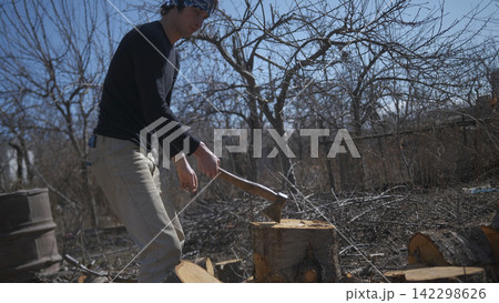 A handsome, brutal man is chopping firewood with an axe. The action takes place in a wooded area with trees and bushes A handsome, brutal man is chopping firewood with an axe. The action takes place in a wooded area with trees and bushes 142298626