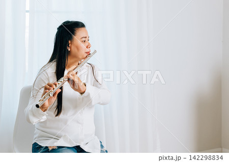 A flutist in a white shirt sits on a chair at home, blowing into her silver flute. The living room curtain backdrop accentuates 142298834