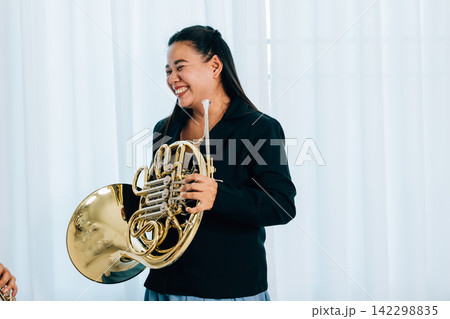 A woman stands at home in front of elegant curtains, playing a shiny gold horn. The warm lighting and her graceful demeanor bring out the beauty of her musical A woman stands at home in front of elegant curtains, playing a shiny gold horn. The warm lighting and her graceful demeanor bring out the beauty of her musical 142298835