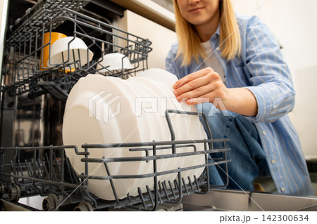 Dishwasher. Woman unloads clean plates from dishwasher in functional kitchen Dishwasher. Woman unloads clean plates from dishwasher in functional kitchen 142300634