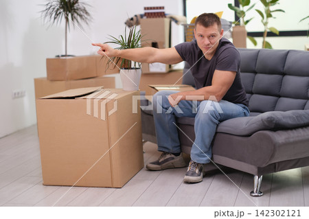 Man points aside with hand as sits on sofa near cardboard box Man points aside with hand as sits on sofa near cardboard box 142302121