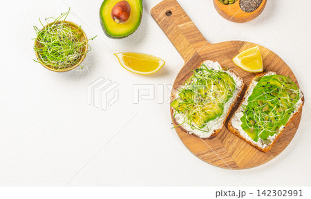 Sliced avocado on whole grain bread topped with sprouts and lemon wedges, presented on a wooden cutting board with additional avocado and seeds in the background 142302991