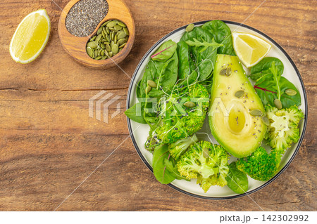 Fresh green salad with avocado, broccoli, spinach, and pumpkin seeds served on a white plate with lime wedges on a wooden table 142302992