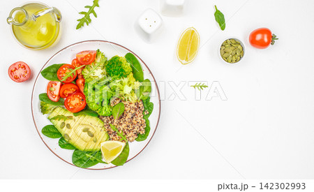 Fresh vegetable salad with quinoa, cherry tomatoes, avocado, and broccoli on a white plate, surrounded by olive oil, lemon, and pumpkin seeds on a clean surface 142302993