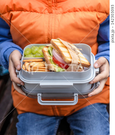 Child holding a gray lunchbox filled with a sandwich, grapes, and crackers, wearing an orange puffer jacket and blue sweater, seated outdoors on a wooden bench Child holding a gray lunchbox filled with a sandwich, grapes, and crackers, wearing an orange puffer jacket and blue sweater, seated outdoors on a wooden bench 142303093