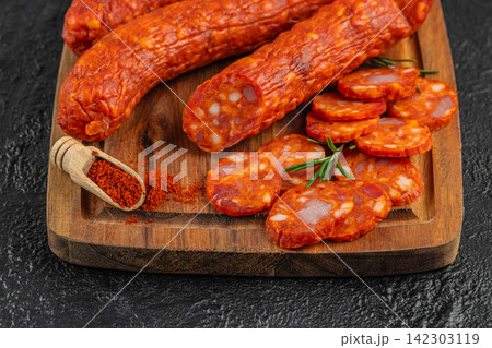 Slices of spicy chorizo sausage arranged on a wooden cutting board with whole chorizo links and a scoop of paprika spice alongside fresh rosemary sprigs 142303119