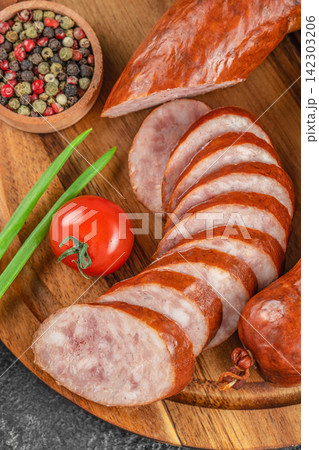 Sliced smoked sausage arranged on a wooden board with fresh tomato, green onions, and a bowl of mixed peppercorns in a rustic kitchen setting 142303206