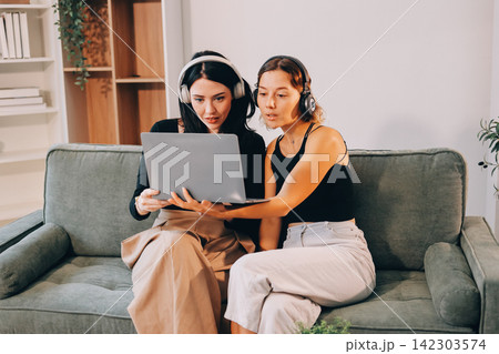 Two young female students using a laptop while sitting on a sofa at home. Student life concept with studying, teamwork, and online learning. 142303574