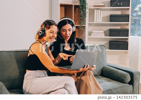 Two young female students using a laptop while sitting on a sofa at home. Student life concept with studying, teamwork, and online learning. Two young female students using a laptop while sitting on a sofa at home. Student life concept with studying, teamwork, and online learning. 142303575