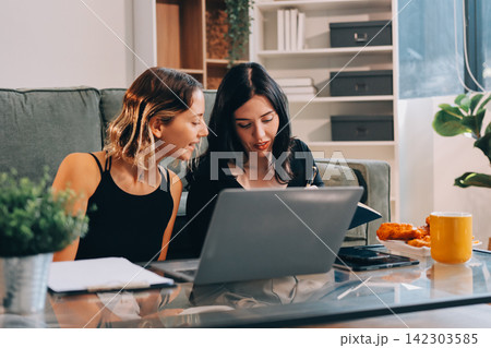 Two young female students using a laptop while sitting on a sofa at home. Student life concept with studying, teamwork, and online learning. 142303585