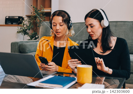 Two young female students using a laptop while sitting on a sofa at home. Student life concept with studying, teamwork, and online learning. Two young female students using a laptop while sitting on a sofa at home. Student life concept with studying, teamwork, and online learning. 142303600