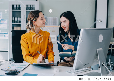 Two businesswomen discussing work using a laptop in a modern office lounge area, showing teamwork and communication Two businesswomen discussing work using a laptop in a modern office lounge area, showing teamwork and communication 142303634