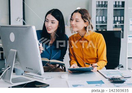 Two businesswomen discussing work using a laptop in a modern office lounge area, showing teamwork and communication 142303635