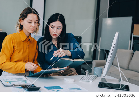 Two businesswomen discussing work using a laptop in a modern office lounge area, showing teamwork and communication 142303636