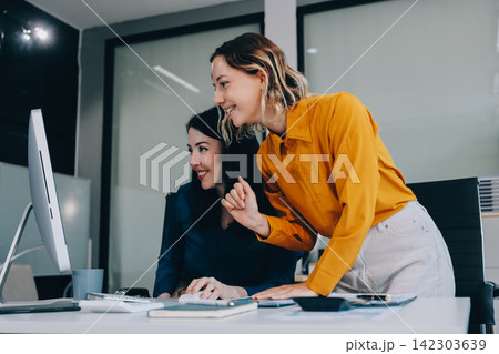 Two businesswomen discussing work using a laptop in a modern office lounge area, showing teamwork and communication Two businesswomen discussing work using a laptop in a modern office lounge area, showing teamwork and communication 142303639
