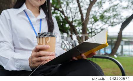 Cropped view of businesswoman holding paper coffee cup and reading notebook outdoors on bench 142303674