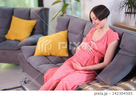 Joyful pregnant woman gently strokes large belly sitting on sofa Joyful pregnant woman gently strokes large belly sitting on sofa 142304385