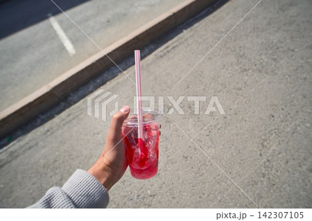hand holding a transparent plastic cup filled with a cold pink berry beverage and ice cubes on a city sidewalk hand holding a transparent plastic cup filled with a cold pink berry beverage and ice cubes on a city sidewalk 142307105