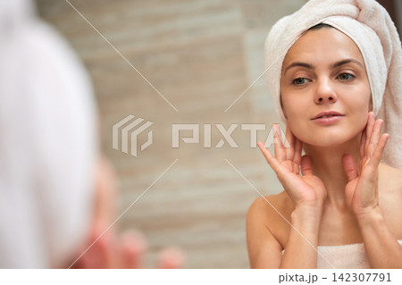 Woman applying skincare, looking at her reflection in mirror Woman applying skincare, looking at her reflection in mirror 142307791