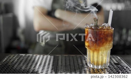 Smoked cocktail with rosemary and ice on a bar, a bartender preparing drinks in background Smoked cocktail with rosemary and ice on a bar, a bartender preparing drinks in background 142308362