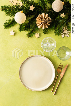 A festive Christmas table setting with an empty plate and cutlery on a bright green background. Spruse branches and golden tree decorations create a warm, holiday atmosphere A festive Christmas table setting with an empty plate and cutlery on a bright green background. Spruse branches and golden tree decorations create a warm, holiday atmosphere 142309504