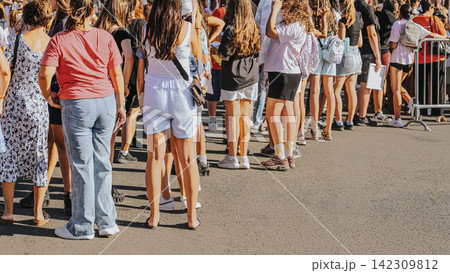 Crowd of young people stands in line. Outdoor Public Event. Large crowd of people standing outdoors at public event on sunny summer day, viewed from behind Crowd of young people stands in line. Outdoor Public Event. Large crowd of people standing outdoors at public event on sunny summer day, viewed from behind 142309812