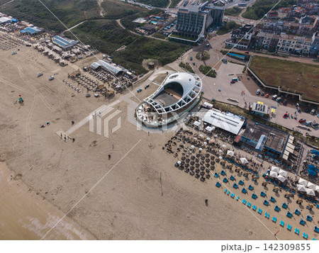 High angle wide horizontal aerial drone photograph of Scheveningen beach promenade, modern beach clubs, curved restaurant, dunes, and coastal skyline in Scheveningen, The Hague, Netherlands High angle wide horizontal aerial drone photograph of Scheveningen beach promenade, modern beach clubs, curved restaurant, dunes, and coastal skyline in Scheveningen, The Hague, Netherlands 142309855