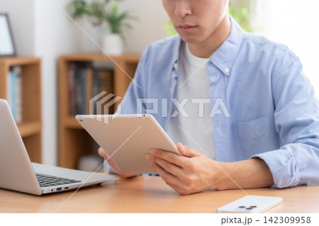 家でノートパソコンとタブレットを見る若い男性 142309958