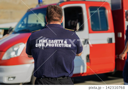 Fire Safety Officer Standing by fire engine. Uniformed fire safety and civil protection officer seen from behind beside red fire engine response vehicle outdoors Fire Safety Officer Standing by fire engine. Uniformed fire safety and civil protection officer seen from behind beside red fire engine response vehicle outdoors 142310768