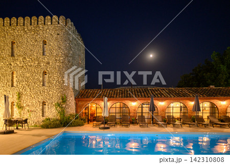 A luxurious hotel resort at night, featuring a grand stone structure with a cylindrical tower and an illuminated outdoor patio 142310808