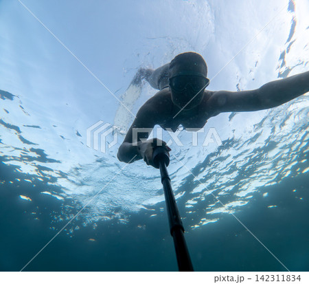 Swimming goggles wearer taking action selfie with monopod while diving in crystal clear sea water 142311834