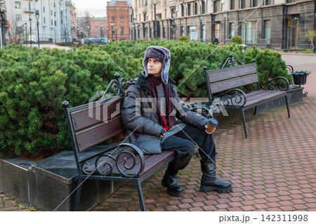 Man student sitting on bench and drinking coffee in city park. Young guy wearing fur hat resting outdoors with hot beverage. Lifestyle leisure in cold weather, urban street scene. Man student sitting on bench and drinking coffee in city park. Young guy wearing fur hat resting outdoors with hot beverage. Lifestyle leisure in cold weather, urban street scene. 142311998