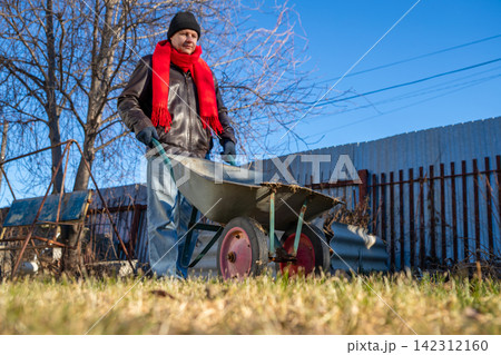 Man pushing wheelbarrow in yard during winter. Adult gardener working with equipment in garden. House maintenance, seasonal yard cleanup and rural lifestyle concept in autumn season. Man pushing wheelbarrow in yard during winter. Adult gardener working with equipment in garden. House maintenance, seasonal yard cleanup and rural lifestyle concept in autumn season. 142312160