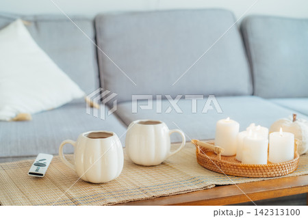 Autumn fall mood composition for hygge home decor. White pumpkin's shaped cups with hot drink, burning candles, remote controller on coffee table in living room. Movie night at home. Cozy relax time. Autumn fall mood composition for hygge home decor. White pumpkin's shaped cups with hot drink, burning candles, remote controller on coffee table in living room. Movie night at home. Cozy relax time. 142313100