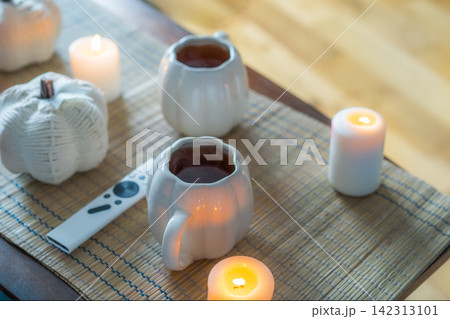 Autumn fall mood composition for hygge home decor. White pumpkin's shaped cups with hot drink, burning candles, remote controller on coffee table in living room. Movie night at home. Cozy relax time Autumn fall mood composition for hygge home decor. White pumpkin's shaped cups with hot drink, burning candles, remote controller on coffee table in living room. Movie night at home. Cozy relax time 142313101